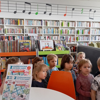 Dzieci w sali w bibliotece. 
Z tyłu regały z książkami.