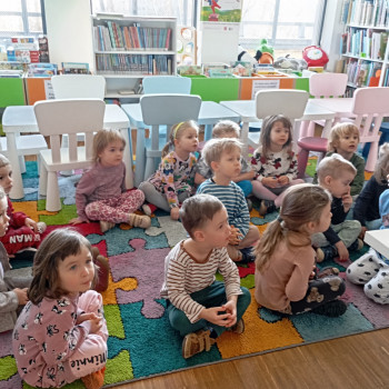 Dzieci siedzą na matach w sali, z tyłu reagały z książkami. 